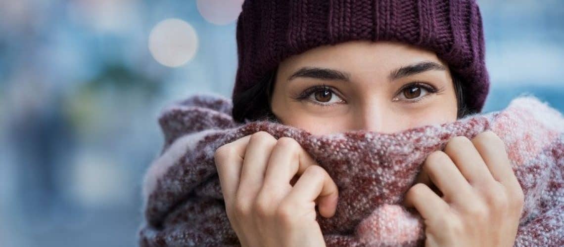 Winter portrait of young beautiful woman covering face with woolen scarf. Closeup of happy girl feeling cold outdoor in the city. Young woman holding scarf and looking at camera. She is smiling and content with her life.