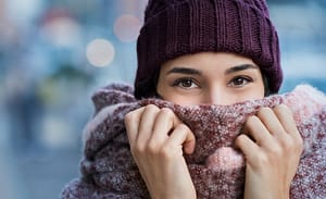 Winter portrait of young beautiful woman covering face with woolen scarf. Closeup of happy girl feeling cold outdoor in the city. Young woman holding scarf and looking at camera. She is smiling and content with her life.