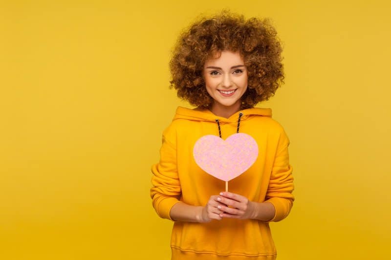An image of a curly haired woman holding a paper heart, indicating kindness makes the world a better place.